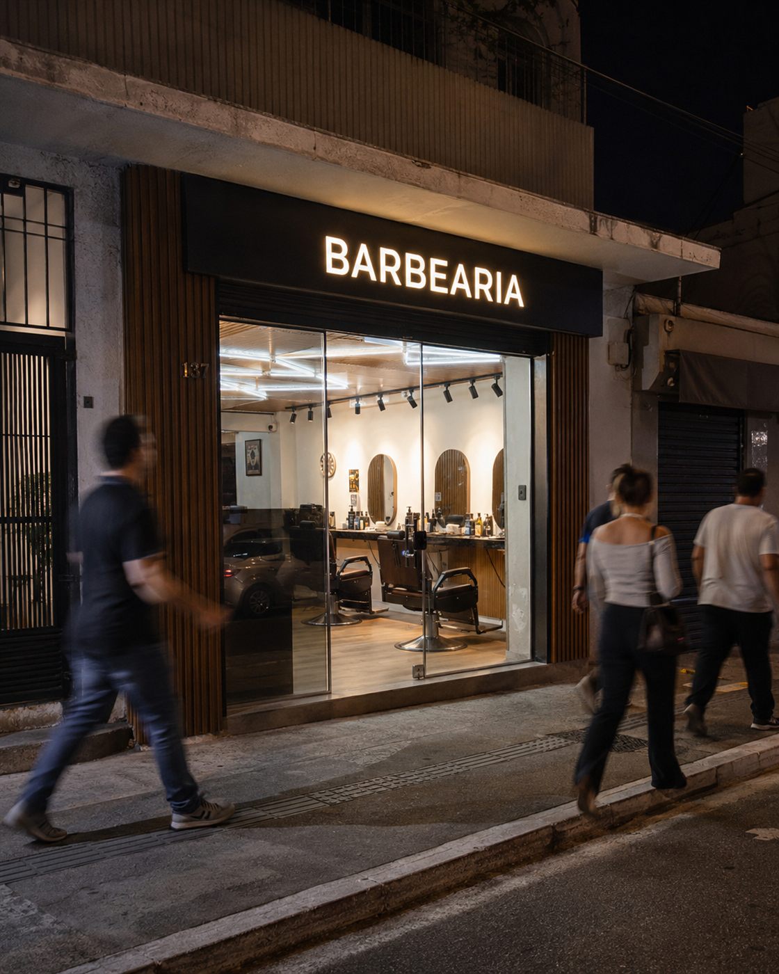 Fachada de barbearia à noite, letreiro aceso e movimento na calçada